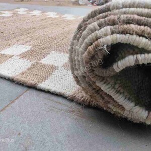 K&ouml;nnte beinhalten: Nahaufnahme eines beige-wei&szlig;en karierten Teppichs mit einem aufgerollten Abschnitt des Teppichs, der die gewebte Textur zeigt. Der Teppich ist aus Naturfasern gefertigt und hat einen rustikalen Look.