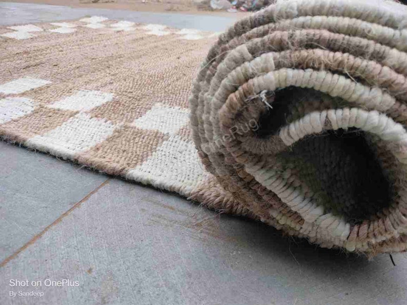 K&ouml;nnte beinhalten: Nahaufnahme eines beige-wei&szlig;en karierten Teppichs mit einem aufgerollten Abschnitt des Teppichs, der die gewebte Textur zeigt. Der Teppich ist aus Naturfasern gefertigt und hat einen rustikalen Look.