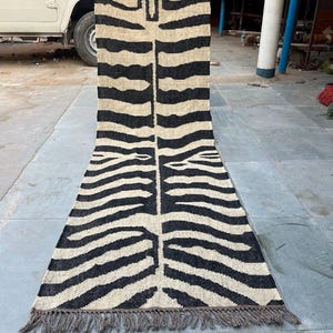 May include: A long, rectangular runner rug featuring a black and beige zebra stripe pattern. The rug has a woven texture with a central vertical line and fringe detailing along the bottom edge. The design displays a stylized animal print motif.