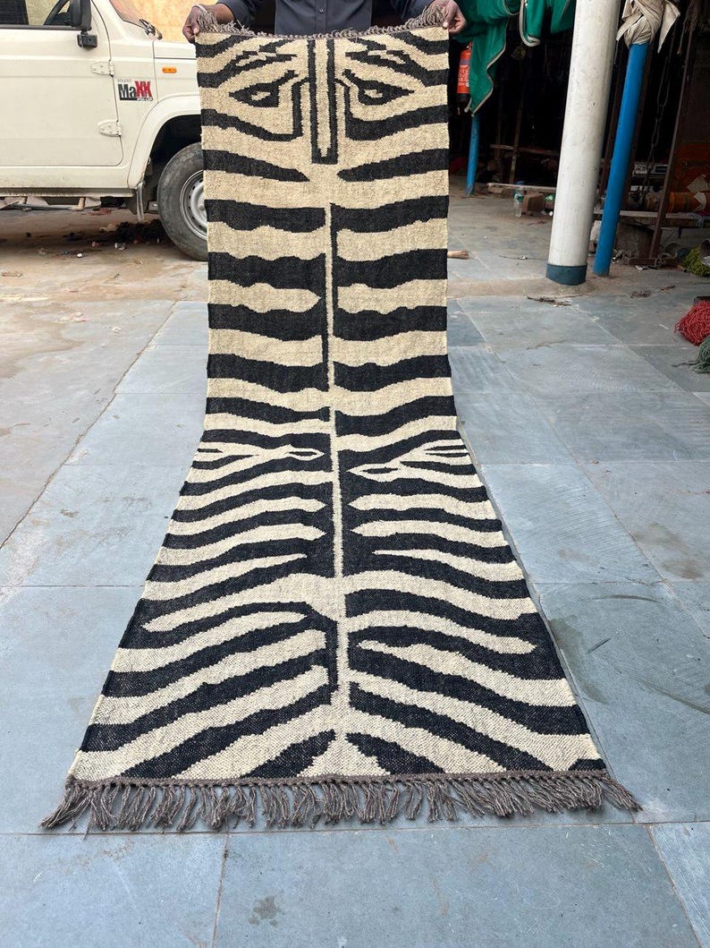 May include: A long, rectangular runner rug featuring a black and beige zebra stripe pattern. The rug has a woven texture with a central vertical line and fringe detailing along the bottom edge. The design displays a stylized animal print motif.