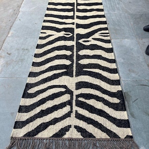 May include: A long, rectangular runner rug featuring a high-contrast black and cream zebra stripe pattern. The woven textile has a symmetrical design running down the center and is finished with short, dark brown fringe tassels on both ends.