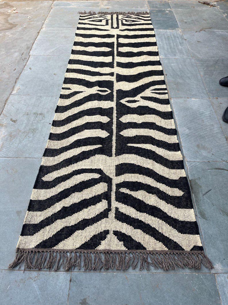 May include: A long, rectangular runner rug featuring a high-contrast black and cream zebra stripe pattern. The woven textile has a symmetrical design running down the center and is finished with short, dark brown fringe tassels on both ends.