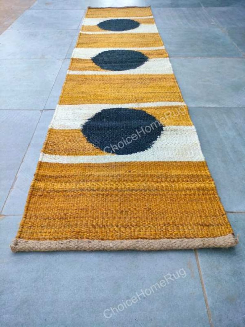 May include: A long, narrow, woven rug with a natural, beige color and black circles. The rug has a white border and is made of natural fibers.