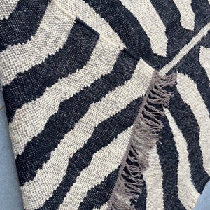 May include: A woven rug featuring a high-contrast black and off-white zebra stripe pattern. The textile has a thick, textured weave and is finished with a row of long, frayed fringe along the edge where the corner is folded over to reveal the back.