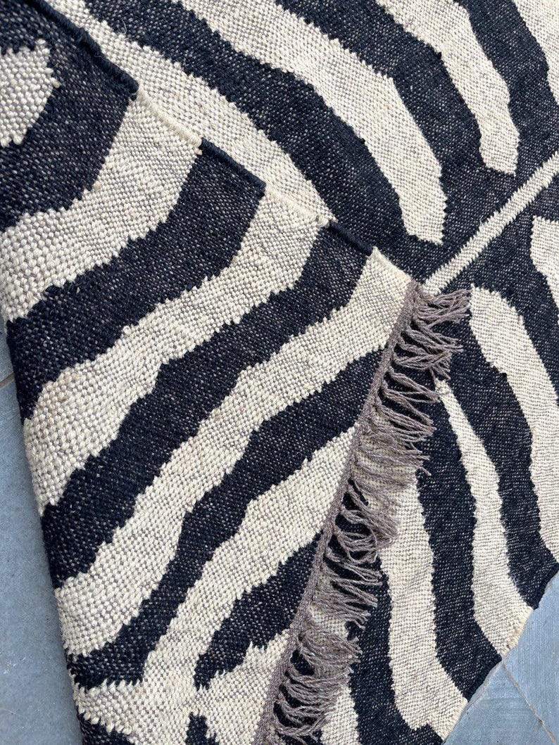 May include: A woven rug featuring a high-contrast black and off-white zebra stripe pattern. The textile has a thick, textured weave and is finished with a row of long, frayed fringe along the edge where the corner is folded over to reveal the back.
