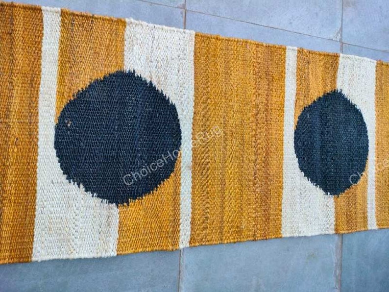 May include: A long, narrow rug with a striped pattern in white, yellow, and black. The rug has two large black circles on it.