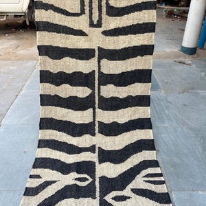 May include: A long, rectangular woven runner rug featuring a high-contrast black and beige zebra stripe pattern. The textile displays a symmetrical design with a central vertical line and horizontal stripes extending outward across the entire surface.
