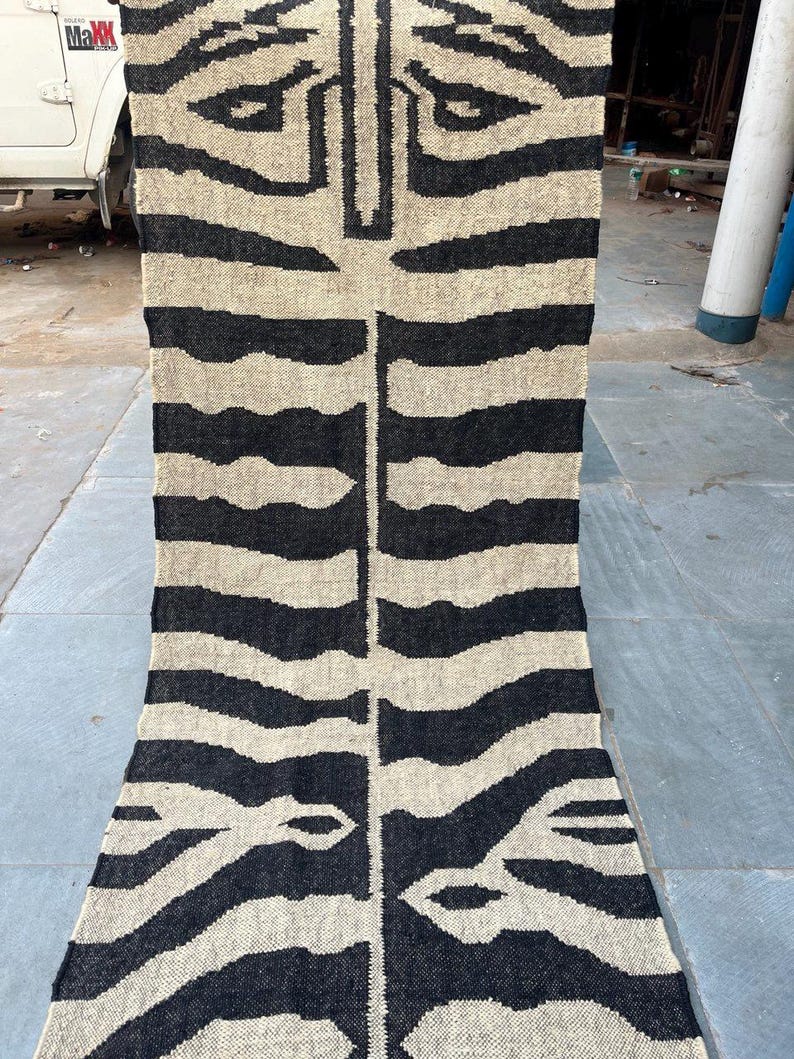 May include: A long, rectangular woven runner rug featuring a high-contrast black and beige zebra stripe pattern. The textile displays a symmetrical design with a central vertical line and horizontal stripes extending outward across the entire surface.