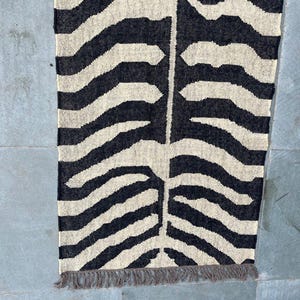 May include: A rectangular woven runner rug featuring a high-contrast black and off-white zebra stripe pattern. The design includes a central vertical seam where the stripes meet, and the bottom edge is finished with short, frayed gray fringe.