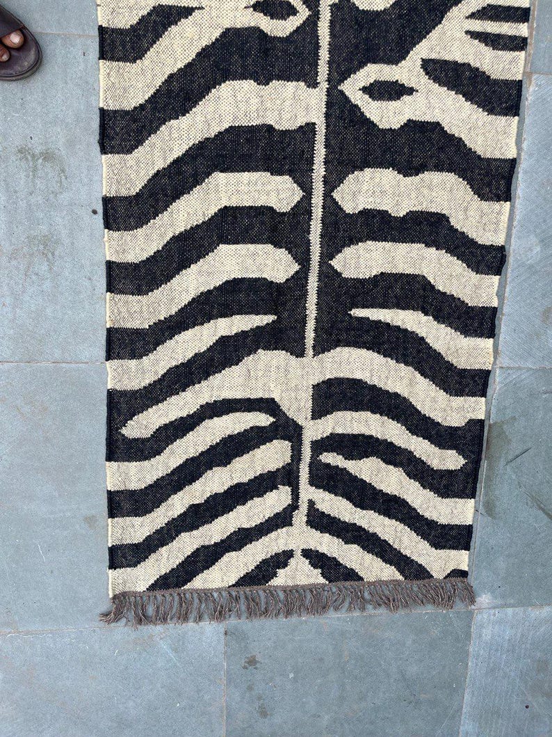 May include: A rectangular woven runner rug featuring a high-contrast black and off-white zebra stripe pattern. The design includes a central vertical seam where the stripes meet, and the bottom edge is finished with short, frayed gray fringe.