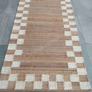 K&ouml;nnte beinhalten: Ein brauner und wei&szlig;er karierter Jute-Teppich mit gewebtem Design. Der Teppich hat ein nat&uuml;rliches, erdiges Aussehen und Gef&uuml;hl.