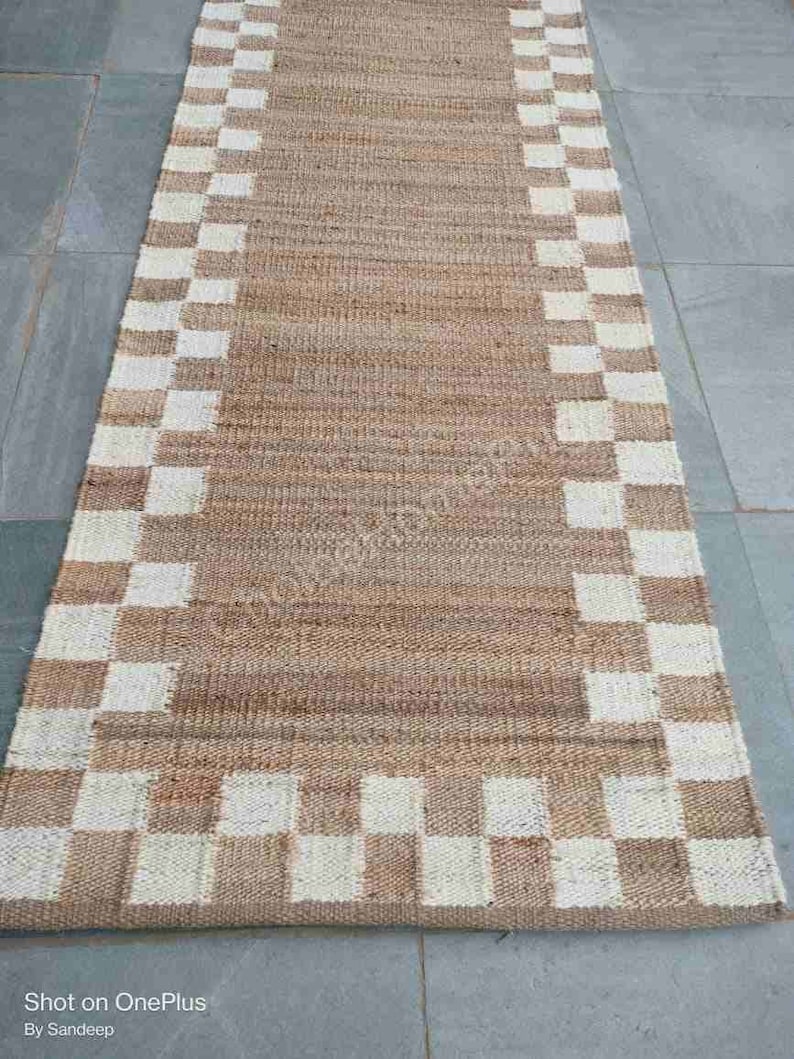 K&ouml;nnte beinhalten: Ein brauner und wei&szlig;er karierter Jute-Teppich mit gewebtem Design. Der Teppich hat ein nat&uuml;rliches, erdiges Aussehen und Gef&uuml;hl.