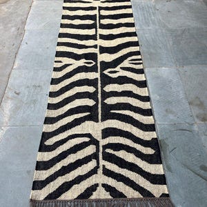 May include: A long, rectangular runner rug featuring a high-contrast black and cream zebra stripe pattern. The woven textile has a symmetrical design running down the center and is finished with short, dark brown fringe along both narrow ends.