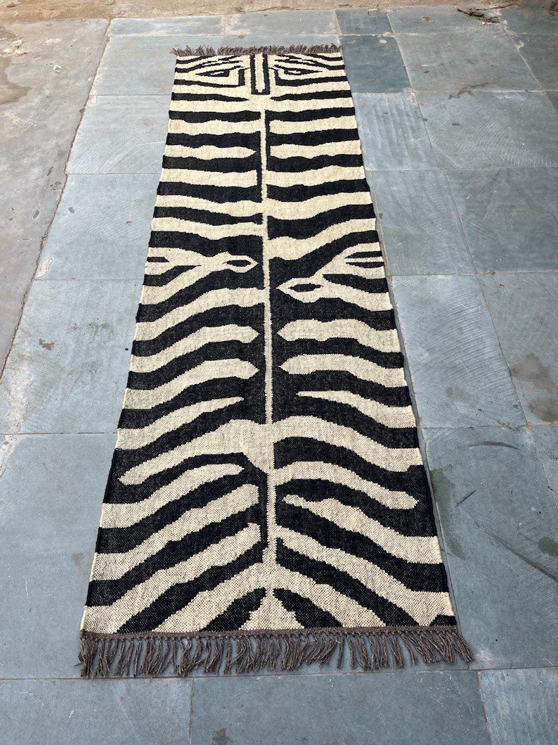 May include: A long, rectangular runner rug featuring a high-contrast black and cream zebra stripe pattern. The woven textile has a symmetrical design running down the center and is finished with short, dark brown fringe along both narrow ends.