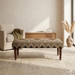 Può includere: Panca rettangolare con piano in tessuto fantasia nei toni del marrone, nero e beige. La panca ha quattro gambe in legno tornito. La panca si trova in una stanza con una palette di colori neutri, una poltrona beige e un mobile in legno.