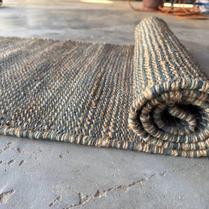 Pode incluir: Um tapete retangular enrolado com uma textura natural e tecida. O tapete apresenta listras alternadas de bege e cinza-azulado. O tapete est&aacute; parcialmente enrolado, revelando o interior enrolado. O tapete &eacute; feito de um material dur&aacute;vel.