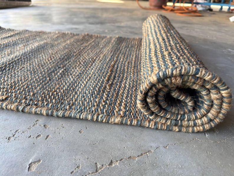 Pode incluir: Um tapete retangular enrolado com uma textura natural e tecida. O tapete apresenta listras alternadas de bege e cinza-azulado. O tapete est&aacute; parcialmente enrolado, revelando o interior enrolado. O tapete &eacute; feito de um material dur&aacute;vel.