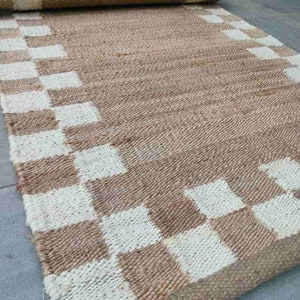 K&ouml;nnte beinhalten: Ein brauner und wei&szlig;er karierter Jute-Teppich mit einem gewebten Design. Der Teppich hat einen Fransenrand und ist aufgerollt.