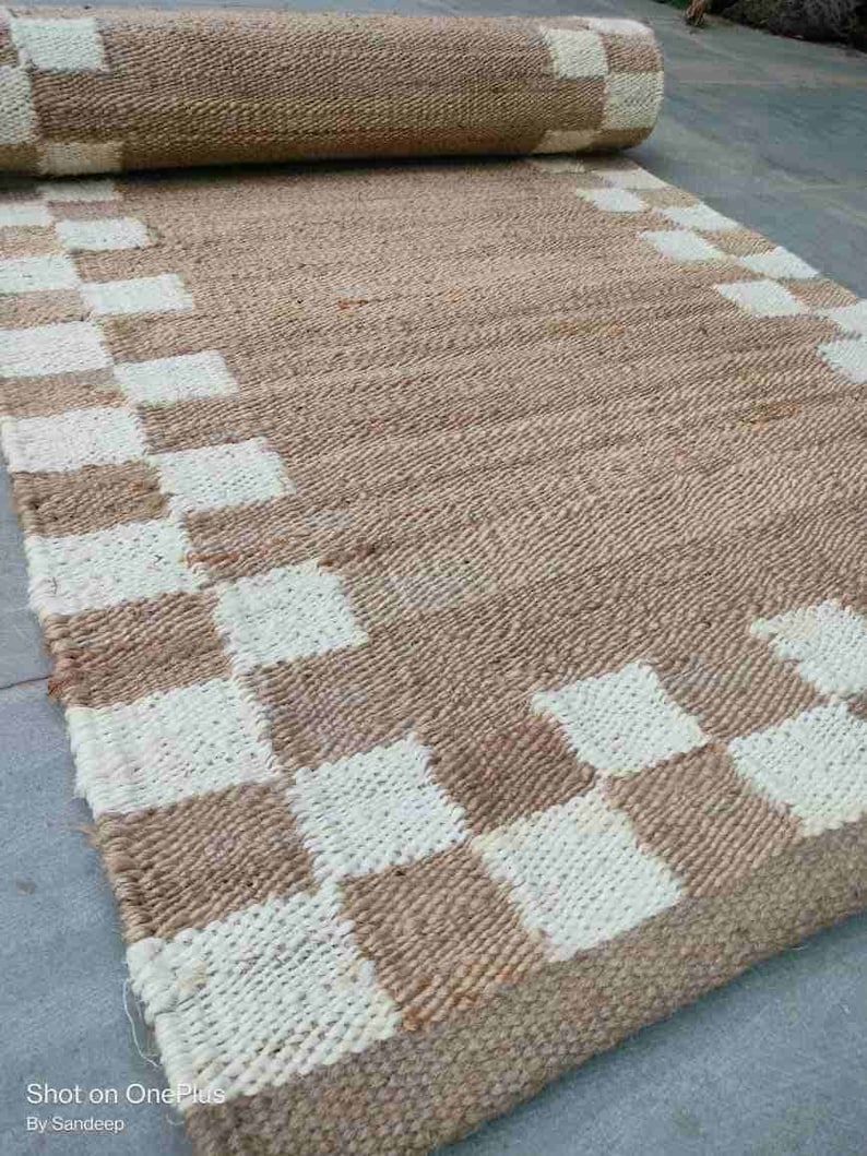 K&ouml;nnte beinhalten: Ein brauner und wei&szlig;er karierter Jute-Teppich mit einem gewebten Design. Der Teppich hat einen Fransenrand und ist aufgerollt.