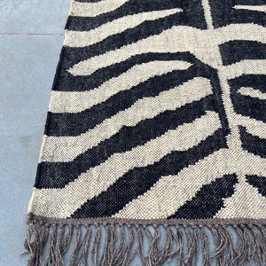 May include: A rectangular woven rug featuring a high-contrast black and cream zebra-inspired striped pattern. The textile has a visible coarse weave texture and is finished with a row of long, loose, light brown fringe along the bottom edge.
