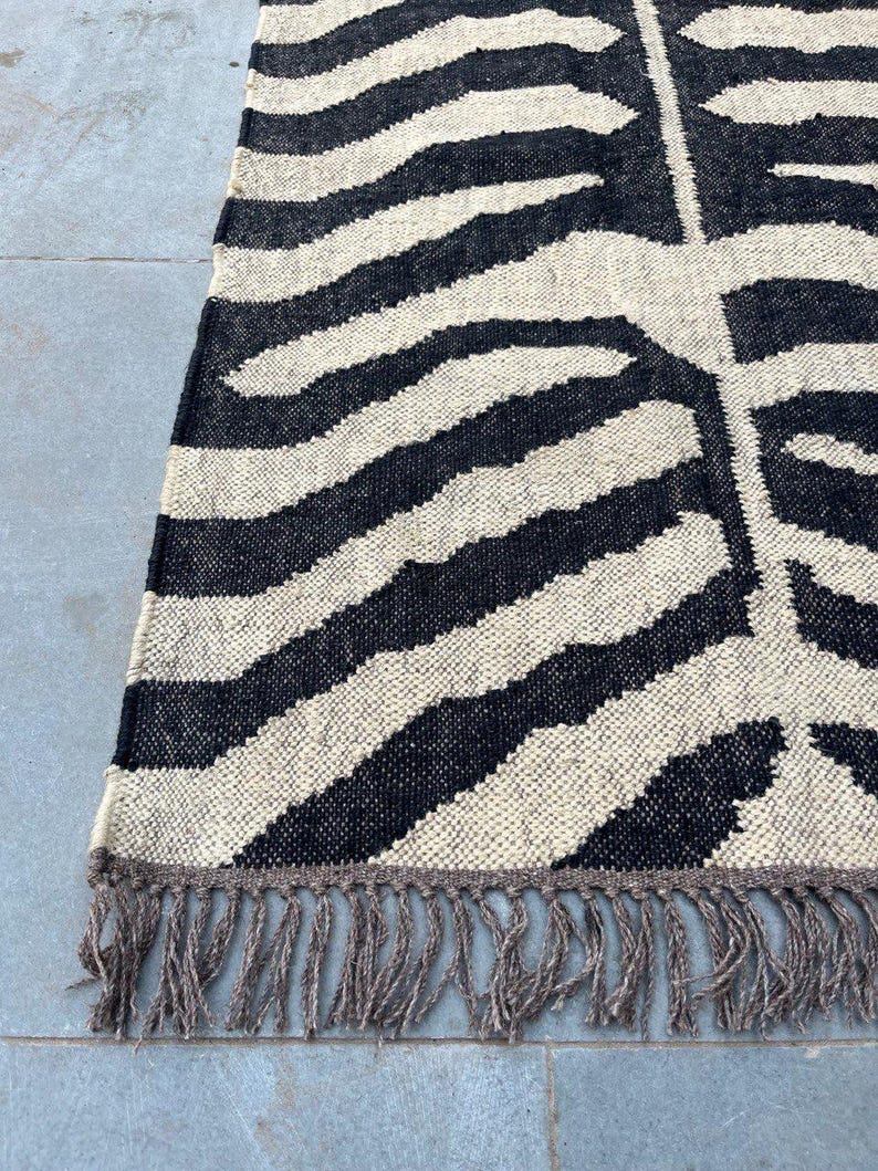 May include: A rectangular woven rug featuring a high-contrast black and cream zebra-inspired striped pattern. The textile has a visible coarse weave texture and is finished with a row of long, loose, light brown fringe along the bottom edge.