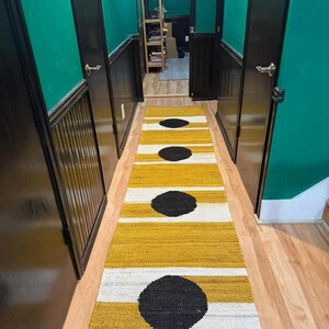 May include: A long, narrow hallway with green walls and black doors. A yellow and white striped rug with black circles runs down the center of the hallway.