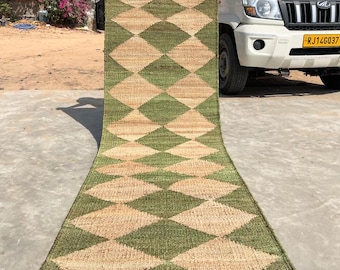 May include: A long, rectangular rug with a diamond pattern in shades of green and beige. The rug is made of natural fibers and has a woven texture.
