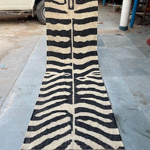 May include: A long, rectangular runner rug featuring a high-contrast black and beige zebra stripe pattern. The woven textile has a central vertical seam and is finished with short, frayed fringe detailing along both the top and bottom edges.