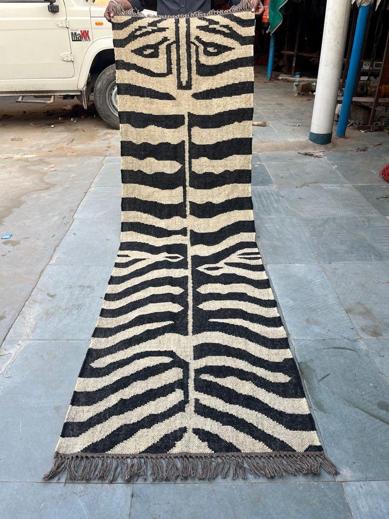 May include: A long, rectangular runner rug featuring a high-contrast black and beige zebra stripe pattern. The woven textile has a central vertical seam and is finished with short, frayed fringe detailing along both the top and bottom edges.