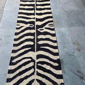 May include: A long, rectangular runner rug featuring a high-contrast black and cream zebra stripe pattern. The woven textile has a symmetrical design running down the center and is finished with short, dark brown fringe along both narrow ends.