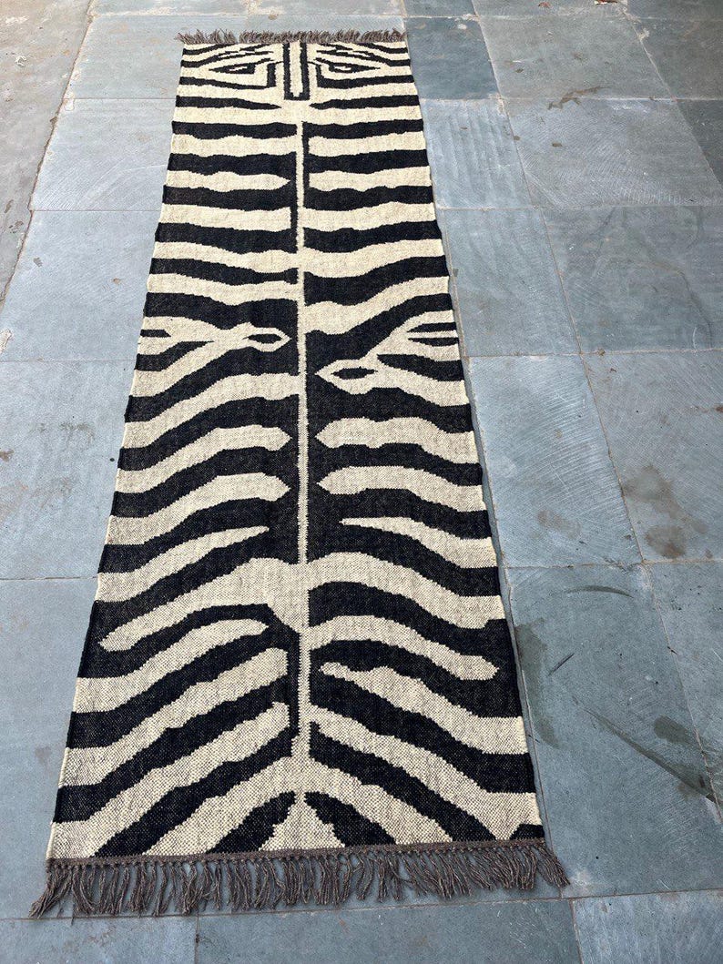 May include: A long, rectangular runner rug featuring a high-contrast black and cream zebra stripe pattern. The woven textile has a symmetrical design running down the center and is finished with short, dark brown fringe along both narrow ends.