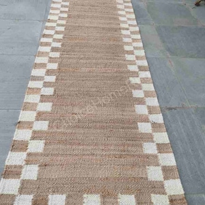 K&ouml;nnte beinhalten: Ein langer, rechteckiger Naturfaser-Teppich mit einem karierten Muster in Wei&szlig; und Braun. Der Teppich hat eine gewebte Textur und ist aus nat&uuml;rlichen Materialien gefertigt.