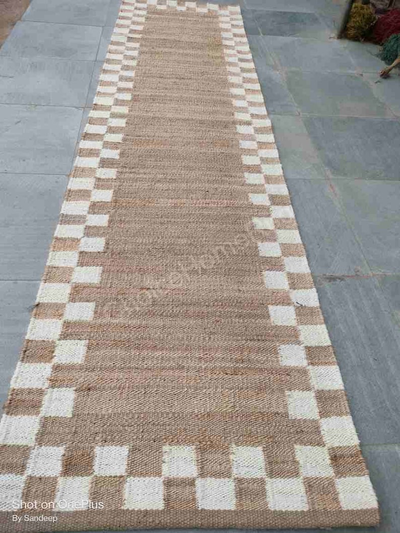 K&ouml;nnte beinhalten: Ein langer, rechteckiger Naturfaser-Teppich mit einem karierten Muster in Wei&szlig; und Braun. Der Teppich hat eine gewebte Textur und ist aus nat&uuml;rlichen Materialien gefertigt.