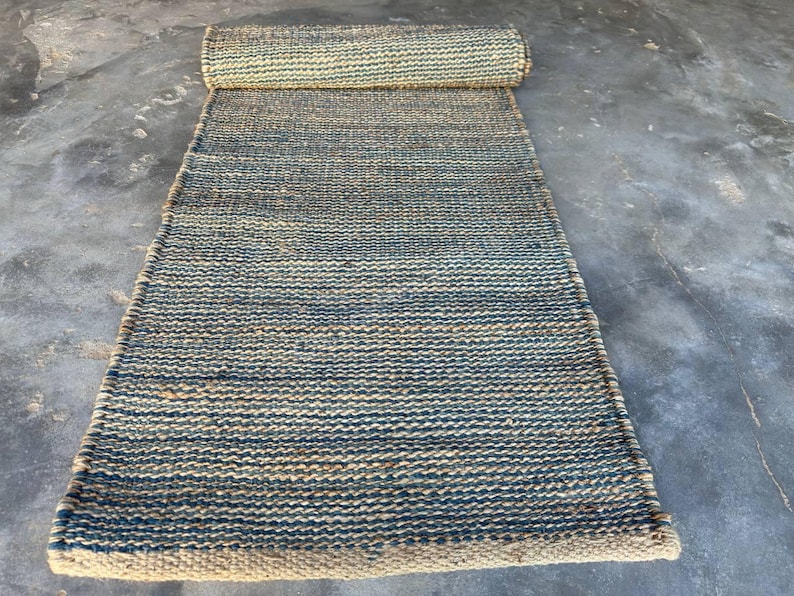 Pode incluir: Um tapete retangular de juta com uma se&ccedil;&atilde;o enrolada na parte superior. O tapete apresenta um design tecido com faixas alternadas de azul e bege natural. O tapete &eacute; adequado para uso interno.