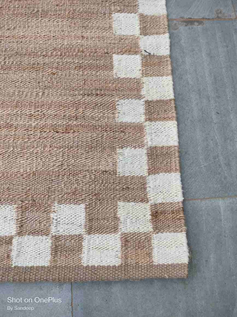 K&ouml;nnte beinhalten: Ein brauner und wei&szlig;er karierter Jute-Teppich mit gewebter Textur. Der Teppich hat einen wei&szlig;en Rand mit einem karierten Muster.