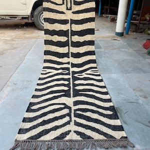 Puede incluir: Una alfombra de pasillo larga y rectangular con un patrón de rayas de cebra en negro y beige de alto contraste. El tejido presenta un diseño simétrico a lo largo del centro y está acabado con flecos cortos deshilachados en ambos extremos.