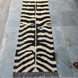 May include: A long, rectangular runner rug featuring a high-contrast black and cream zebra stripe pattern. The woven textile has a symmetrical design running down the center and is finished with short, dark brown fringe tassels on both ends.