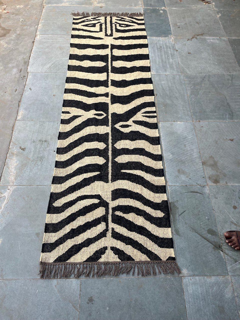 May include: A long, rectangular runner rug featuring a high-contrast black and cream zebra stripe pattern. The woven textile has a symmetrical design running down the center and is finished with short, dark brown fringe tassels on both ends.