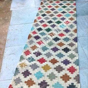 Op de afbeelding: Een lang, rechthoekig loperkleed met een crèmekleurige basis en een herhalend patroon van kleine, veelkleurige kruisvormige motieven in tinten rood, blauw, groen, geel, bruin en grijs, afgewerkt met een franjerand aan de onderkant.