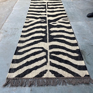 May include: A long rectangular runner rug featuring a high-contrast black and off-white zebra stripe pattern. The woven textile has a symmetrical design running down the center and is finished with short, dark brown fringe along both ends.