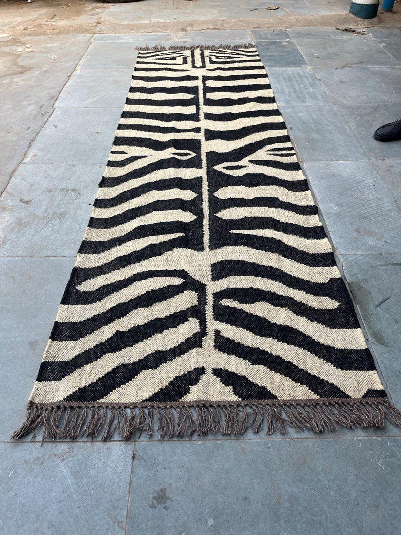 May include: A long rectangular runner rug featuring a high-contrast black and off-white zebra stripe pattern. The woven textile has a symmetrical design running down the center and is finished with short, dark brown fringe along both ends.