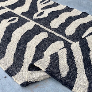 May include: A rectangular woven rug featuring a high-contrast black and off-white zebra-inspired striped pattern. The textile has a thick, textured weave with visible fibers and a folded corner that reveals the underside of the material.
