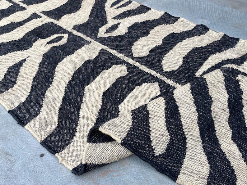 May include: A rectangular woven rug featuring a high-contrast black and off-white zebra-inspired striped pattern. The textile has a thick, textured weave with visible fibers and a folded corner that reveals the underside of the material.