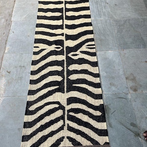 May include: A long, rectangular runner rug featuring a high-contrast black and cream zebra stripe pattern. The woven textile has a symmetrical design running down the center and is finished with short, dark brown fringe along both narrow ends.