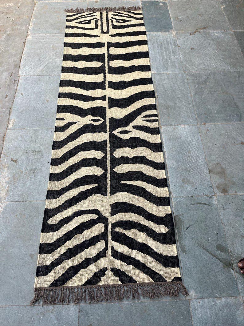 May include: A long, rectangular runner rug featuring a high-contrast black and cream zebra stripe pattern. The woven textile has a symmetrical design running down the center and is finished with short, dark brown fringe along both narrow ends.