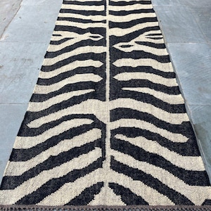 May include: A long, rectangular runner rug featuring a high-contrast black and cream zebra stripe pattern. The woven textile has a textured surface and is finished with short, knotted fringe detailing along both of the narrow ends of the rug.
