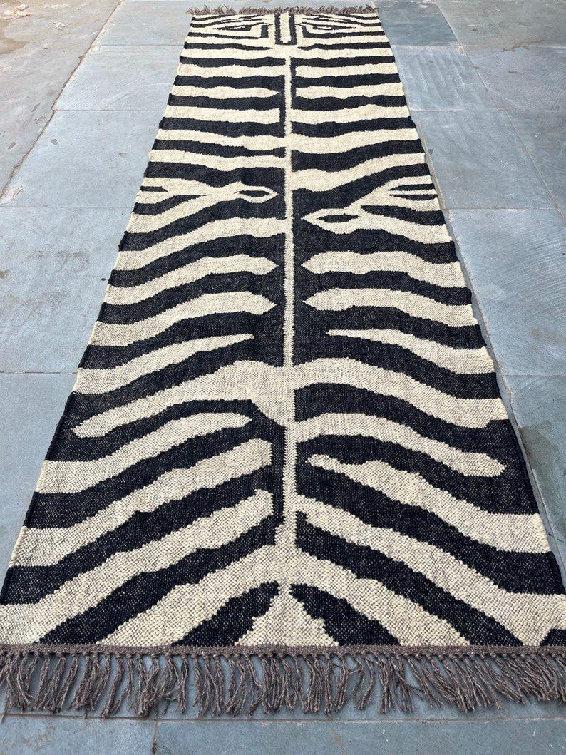 May include: A long, rectangular runner rug featuring a high-contrast black and cream zebra stripe pattern. The woven textile has a textured surface and is finished with short, knotted fringe detailing along both of the narrow ends of the rug.