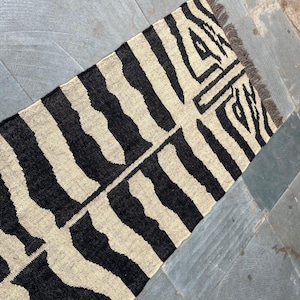 May include: A rectangular woven rug featuring a high-contrast black and off-white zebra-inspired striped pattern. The textile has a visible textured weave and includes short, dark-colored fringe detailing along one of the shorter edges.