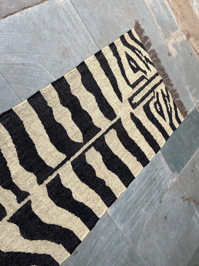 May include: A rectangular woven rug featuring a high-contrast black and off-white zebra-inspired striped pattern. The textile has a visible textured weave and includes short, dark-colored fringe detailing along one of the shorter edges.