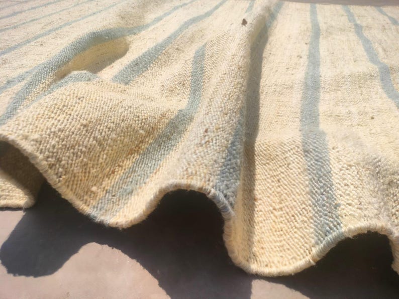 May include: A close-up of a cream-colored rug with blue stripes. The rug is woven and has a textured surface.